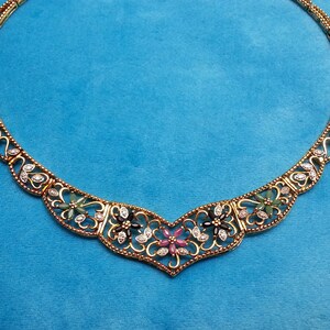 May include: A gold-toned metal necklace featuring an intricate filigree design with a central floral motif. The piece is adorned with small, clear, sparkling stones and inlaid with green, black, and pink petal-shaped gemstones in a symmetrical pattern.