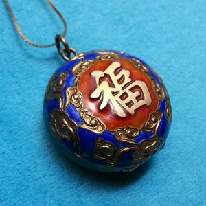 May include: A round cloisonné pendant on a thin metal chain. The pendant features a central orange enamel circle with a raised silver-toned Chinese character for good fortune, surrounded by a deep blue enamel border with gold-toned cloud motifs.