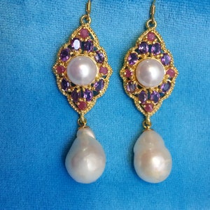 May include: A pair of gold-tone dangle earrings featuring a central white pearl surrounded by small purple and red stones in a diamond-shaped setting. A large, irregular white baroque pearl hangs from the bottom of each ornate gold-tone metal frame.