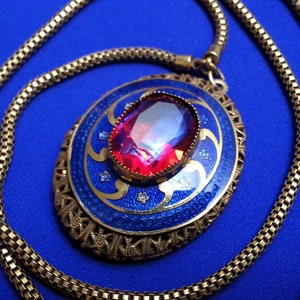 May include: An oval pendant necklace featuring a large, faceted, deep pink gemstone set in the center. The pendant has a blue enamel background with gold-toned swirling patterns and small clear stones. It hangs from a gold-toned mesh chain.