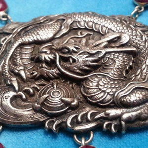 Puede incluir: Collar con colgante plateado con un diseño detallado de dragón. El colgante cuadrado presenta un dragón. El collar tiene detalles de piedras preciosas rojas y se muestra sobre un fondo azul.