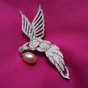 May include: A silver-toned bird brooch encrusted with numerous clear, sparkling stones. The bird is depicted in flight with wings extended upward and a long, curved tail. A single, smooth, oval-shaped light pink pearl hangs from the bird's beak.