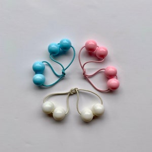 Könnte beinhalten: Drei Haargummis mit doppelten Kugeln. Ein Haargummi ist blau mit zwei hellblauen Kugeln, eines ist rosa mit zwei hellrosa Kugeln und eines ist weiß mit zwei weißen Kugeln.