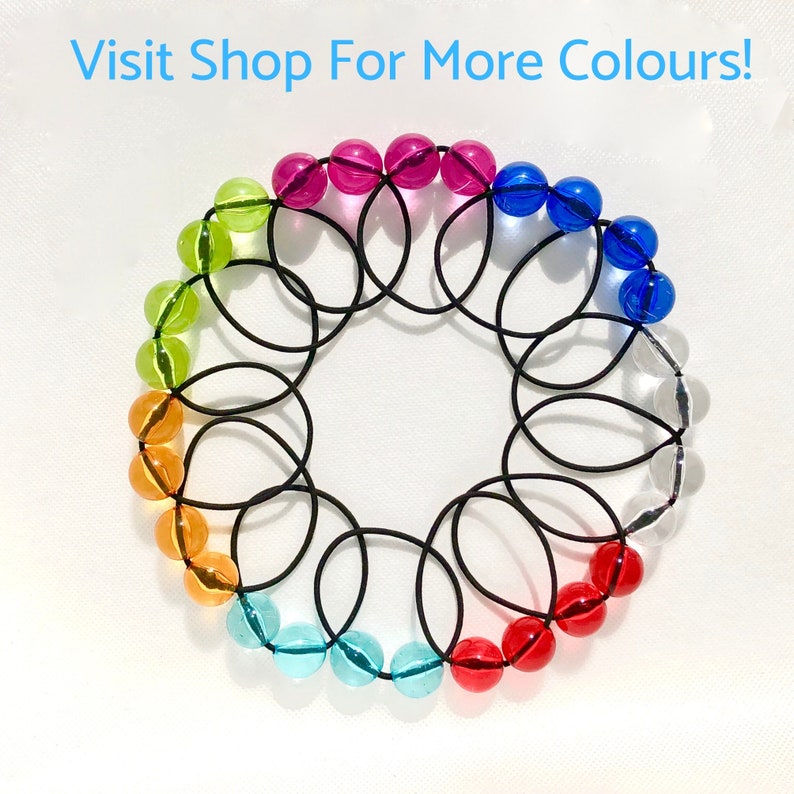 May include: A rainbow of colourful glass beads are strung together on a black elastic band. The beads are arranged in a circle, with the colours transitioning from green to orange, red, blue, and clear. The text "Visit Shop For More Colours!" is written in blue letters at the top of the image.