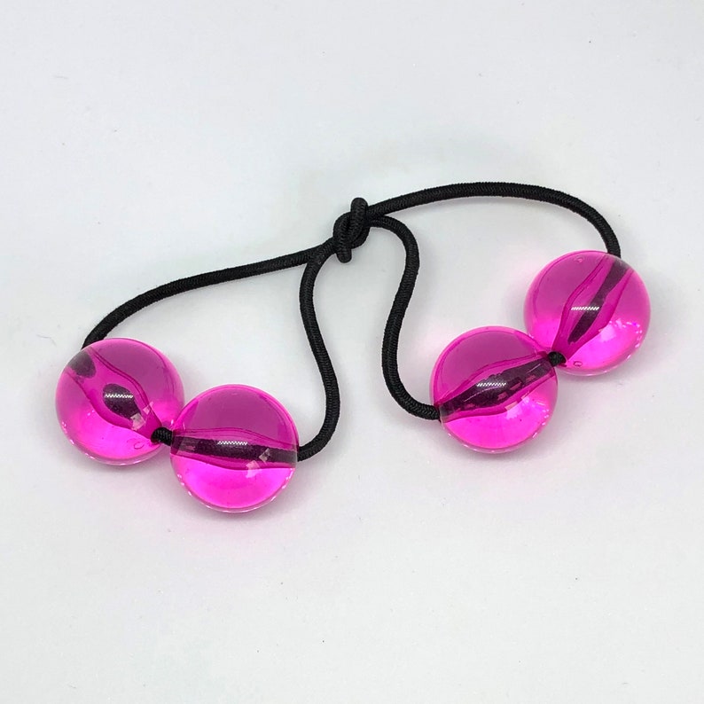 May include: Three pink, clear, round balls connected by a black elastic band. The balls are translucent and have a slight swirl pattern.