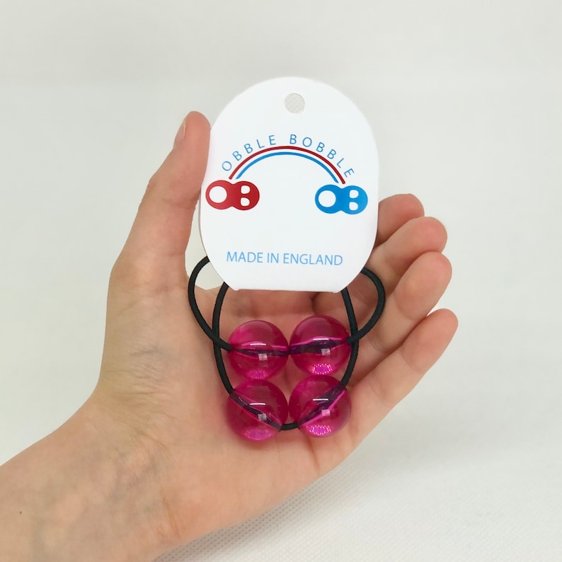 May include: A set of four pink bobble hair ties with a black elastic band. The hair ties are packaged with a white tag that reads "OBBLE BOBBLE" with a rainbow arch and the text "MADE IN ENGLAND".