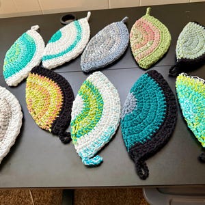 May include: A collection of eleven handmade crochet leaf-shaped pot holders in various colors. Each pot holder features a loop for hanging and is made with a textured stitch. Colors include teal, white, gray, black, yellow, and green.