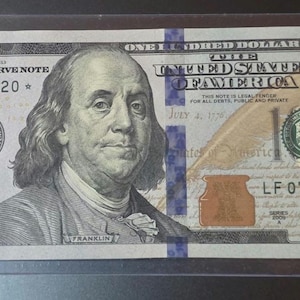 May include: A United States one hundred dollar bill, featuring a portrait of Benjamin Franklin. The bill is encased in a clear protective sleeve. The text "ONE HUNDRED DOLLARS" and "FEDERAL RESERVE NOTE" are visible.