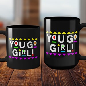 May include: Two black coffee mugs with a white "You Go Girl!" graphic in colorful letters and a rainbow-colored triangle pattern.
