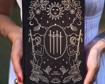 Op de afbeelding: Zwart tarot werkboek met goudfolie illustraties. De cover toont een zon, maan, vlinders en twee figuren met staven. Het midden toont drie zwaarden. De woorden "tarot workbook" staan onderaan gedrukt.