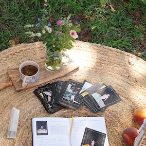 May include: A black and gold tarot deck spread out on a woven rug with a journal open to the Three of Wands card. The journal has handwritten notes about the card's meaning. There is a cup of tea, a crystal, and a peach on the rug.