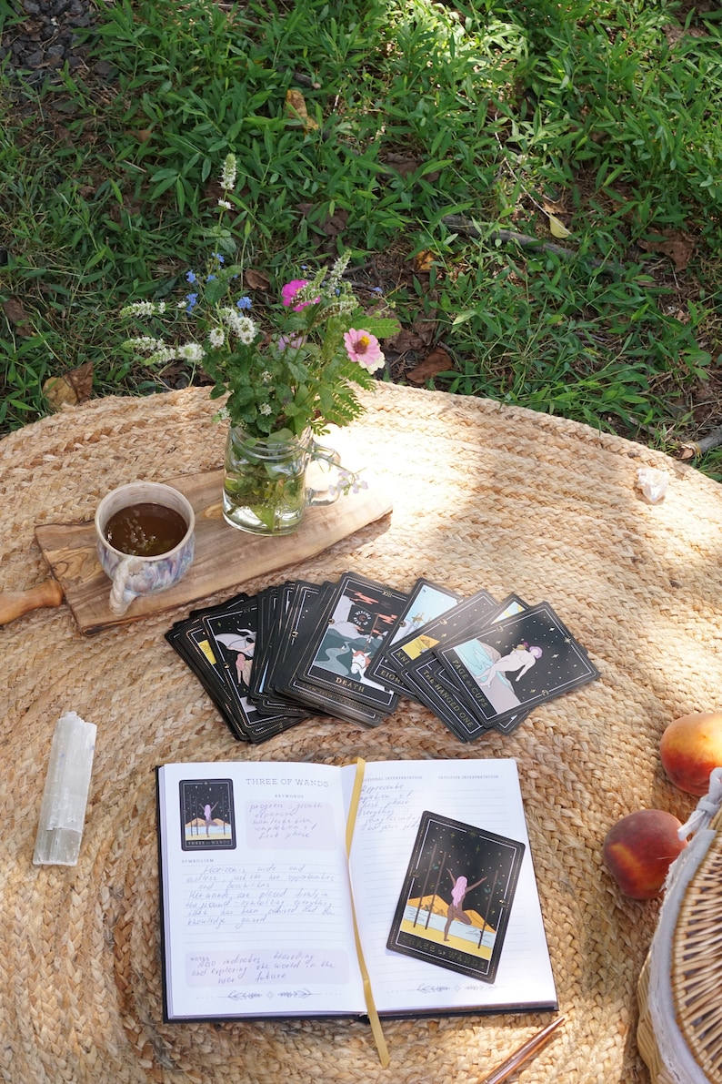 May include: A black and gold tarot deck spread out on a woven rug with a journal open to the Three of Wands card. The journal has handwritten notes about the card's meaning. There is a cup of tea, a crystal, and a peach on the rug.