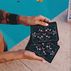 May include: Three black tarot cards with a floral and crescent moon design. The cards are held in the hands of a person.