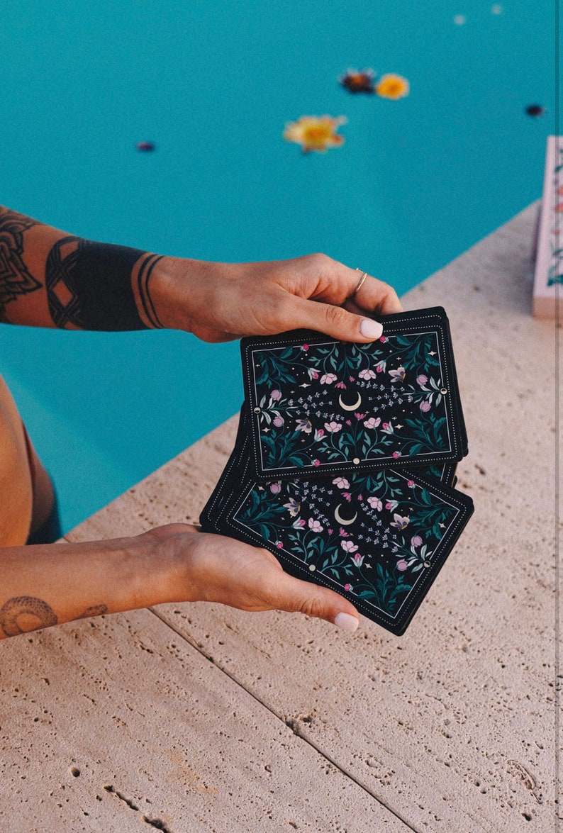 May include: Three black tarot cards with a floral and crescent moon design. The cards are held in the hands of a person.