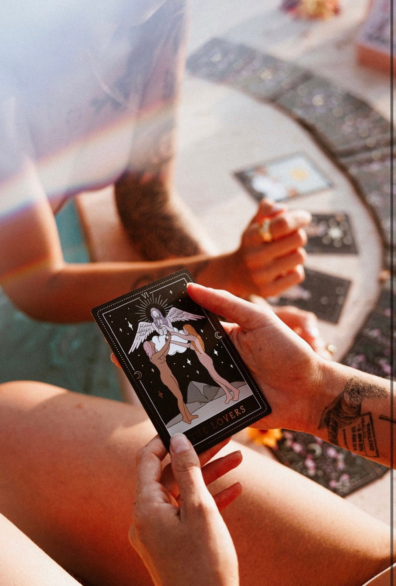 May include: A black tarot card with gold accents depicting the Lovers card. The card shows two figures holding hands and looking up at a winged figure above them. The card is labeled "VII The Lovers".