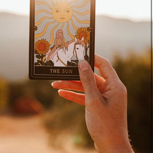 May include: A tarot card with a gold border and the words "THE SUN" printed on the bottom. The card depicts a person riding a white horse with a sun with a face in the background.
