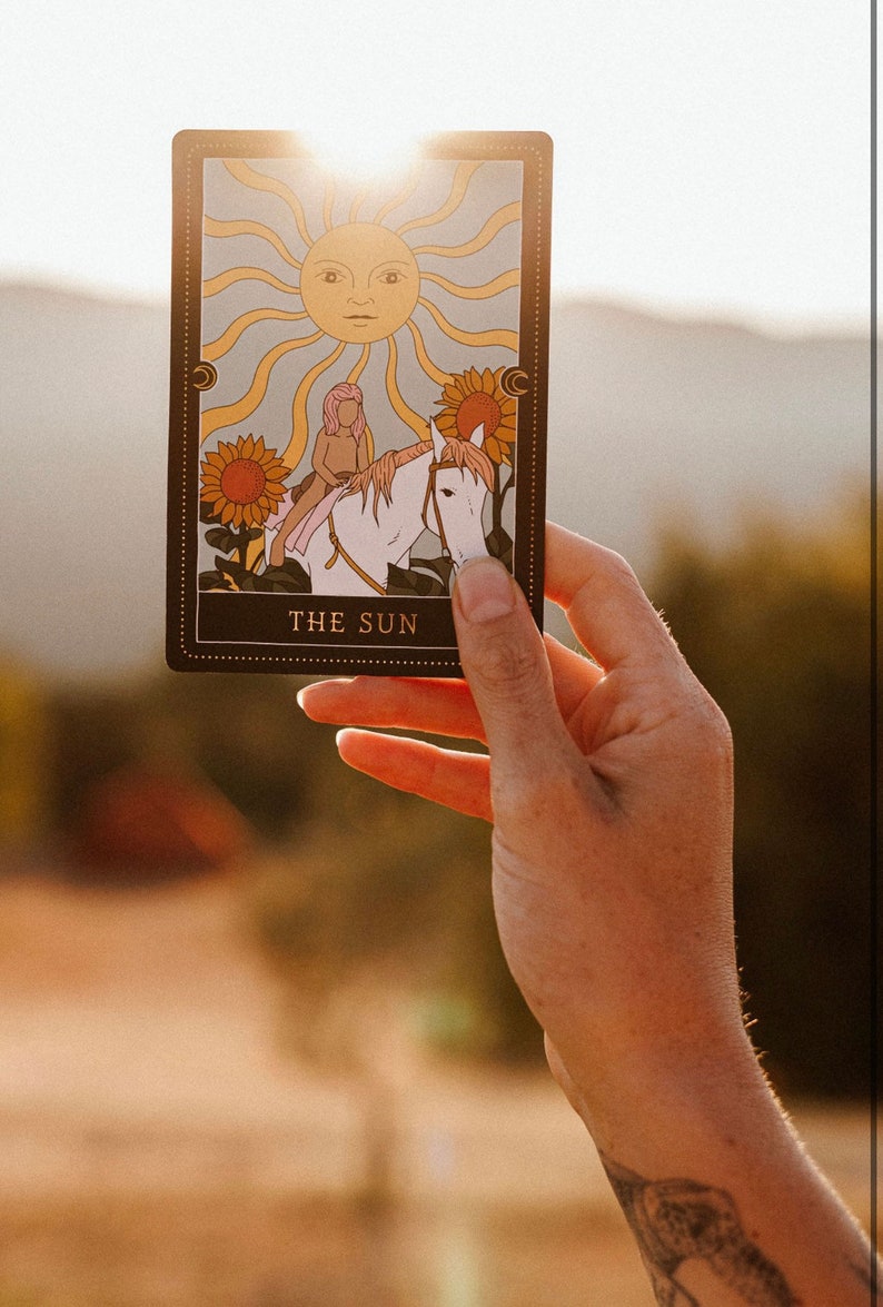 May include: A tarot card with a gold border and the words "THE SUN" printed on the bottom. The card depicts a person riding a white horse with a sun with a face in the background.