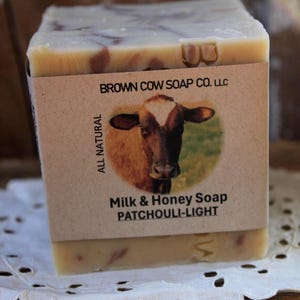May include: A rectangular bar of soap with a light brown and cream color scheme. The soap is wrapped with a label featuring a cow illustration and the text "Milk & Honey Soap PATCHOULI-LIGHT". The words "ALL NATURAL" are also visible.