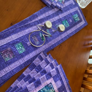 May include: A quilted table runner and matching placemats in shades of purple and lavender. The runner features a rectangular design with square accents of green and blue patterned fabric. A silver candle holder with two white candles sits on the runner.