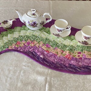 May include: A paisley-shaped table runner in purple, green, and pink, with a tea set. The tea set includes a teapot, creamer, sugar bowl, and two teacups and saucers, all decorated with floral patterns. The runner is made of quilted fabric.