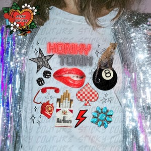 K&ouml;nnte beinhalten: Hellblaues T-Shirt mit einer Collage von Grafiken, darunter rote Lippen, ein Leopard auf einer 8-Kugel, ein Stern, W&uuml;rfel, ein Telefon, eine Packung Marlboro-Zigaretten, ein T&uuml;rkis-Cluster und die Worte "HONKY TONK".