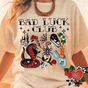 May include: A beige t-shirt with the text "Bad Luck Club" in black lettering. The shirt features a variety of colorful illustrations including a snake, a scorpion, a lighter, a coffin, dice, and flowers.
