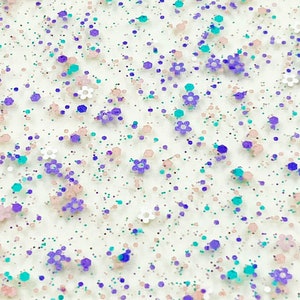 May include: White background with a scattered pattern of small purple, pink, and turquoise glitter. The glitter is in the shape of dots and small flowers.