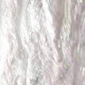 Puede incluir: Primer plano de una superficie blanca, esponjosa y texturizada. El material tiene un aspecto suave, similar a una nube, con sutiles tonos rosados. Pequeñas partículas de purpurina brillante están dispersas por la superficie, añadiendo un toque de brillo.