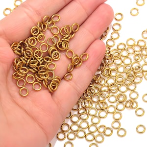May include: A collection of small, gold-colored jump rings, scattered across a hand and a white surface. The rings are circular and appear to be made of metal. They are commonly used in jewelry making and crafting.
