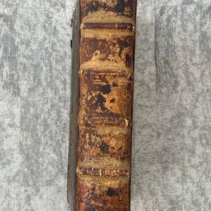 May include: The spine of an old, leather-bound book with a worn, brown cover. The book has raised bands and is showing signs of age and use.