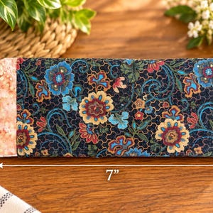 May include: A rectangular fabric case featuring a dark navy blue background with a multi-colored floral pattern in shades of blue, red, and yellow, accented by a light pink textured side panel. The case measures 3.5 inches by 7 inches.