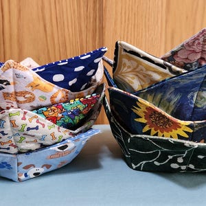 May include: Two stacks of fabric bowl cozies featuring various patterns. The left stack includes blue polka dots, dog-themed prints, and bone motifs. The right stack features floral designs, a dark blue sunflower pattern, and a dark green leaf print.
