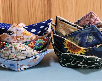 May include: Two stacks of fabric bowl cozies. The left stack features patterns of blue polka dots, cats, colorful dog bones, and cartoon dogs. The right stack displays floral, dark blue sunflower, and dark green leaf patterns.