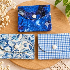 May include: Three small fabric pouches with snap closures. One pouch features a blue galaxy print with stars, one has a blue and white floral pattern, and the third displays a blue and white gingham check design. Each pouch is rectangular.