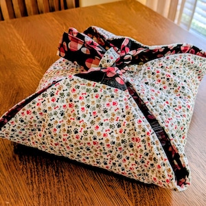 May include: A square fabric gift wrap, featuring a white background with paw prints and bones in red, brown, and gray. The wrap is tied with a black and red patterned bow, creating a decorative and reusable packaging option.