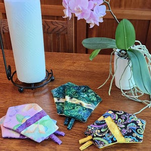May include: A collection of colorful, hexagonal fabric coasters with various patterns, including butterflies and floral designs. The coasters are in shades of purple, green, and black, with yellow accents. A roll of paper towels and a potted orchid are also visible.