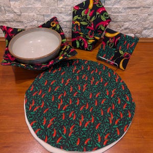 May include: A set of kitchen textiles featuring a black background with a red chili pepper and green leaf pattern. The set includes a bowl cozy, a pot holder, a square coaster, and a round placemat.