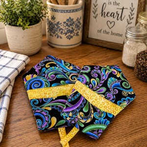 May include: Two black oven mitts with a colorful paisley pattern in blue, purple, and green, accented with yellow bands. The mitts are placed on a wooden surface, suggesting a kitchen setting.