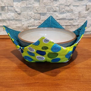 May include: A ceramic bowl rests inside a microwave bowl cozy. The cozy is made of a bright yellow fabric with blue and white spiral designs. The interior and the top triangle are teal with a small dot pattern. The cozy is on a wooden surface.