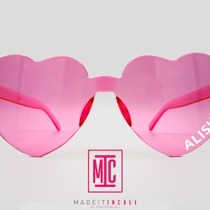 May include: Pink heart-shaped sunglasses with the name "ALISHA" printed on the right lens. The sunglasses are made by MADEITINCALI.