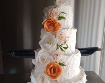 Puede incluir: Un pastel de bodas blanco de cuatro pisos decorado con glaseado de mantequilla y flores de papel. Las flores son una mezcla de blanco y naranja.