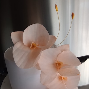 Puede incluir: Un pastel blanco decorado con delicadas flores de orqu&iacute;dea de color melocot&oacute;n. El pastel est&aacute; sobre un soporte de pastel blanco.