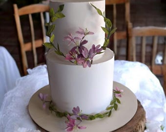 Puede incluir: Pastel de boda blanco de dos niveles decorado con flores moradas y rosas y hojas verdes. El pastel está sobre un soporte para pastel de madera y un mantel de encaje blanco.