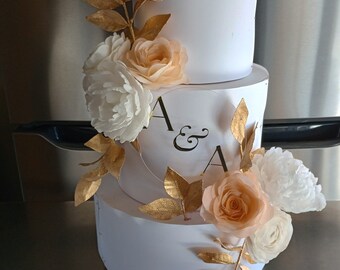 Puede incluir: Un pastel blanco de tres pisos decorado con pan de oro y flores de papel de melocotón y blanco. El pastel está decorado con las iniciales "A & A" en escritura negra.