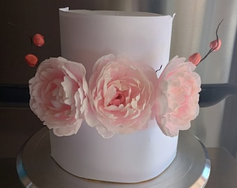 Op de afbeelding: Een witte taart met roze papieren bloemen en rode bessen op een zilveren taartplateau.