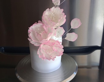 May include: A white cake with pink sugar flowers on a silver cake stand. The flowers are arranged in a cascading design.