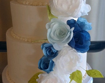 以下が含まれることがあります： 白い3段のウェディングケーキ。白い紙の花、青い紙の花、薄い青色の紙の花、緑の葉で飾られています。