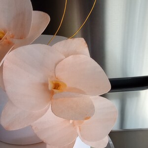 Puede incluir: Un pastel blanco decorado con delicadas flores de orqu&iacute;dea de color melocot&oacute;n. El pastel est&aacute; acentuado con alambre dorado.