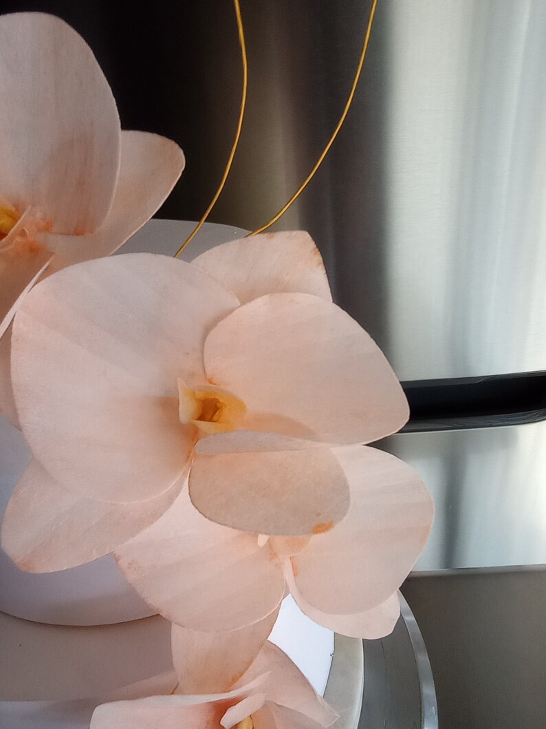 Puede incluir: Un pastel blanco decorado con delicadas flores de orqu&iacute;dea de color melocot&oacute;n. El pastel est&aacute; acentuado con alambre dorado.