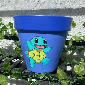 May include: A blue ceramic planter with a hand-painted image of Squirtle, a Pokemon character, in a playful pose. The planter is perfect for adding a touch of whimsy to your home decor.